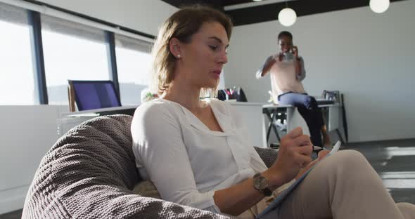 Caucasian businesswoman sitting in armchair, writing while her coworker having business call alt