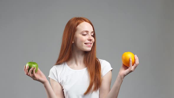 Beautiful Redhead Girl Tries To Choose Orange or Apple. Healthy Food Selection Concept alt