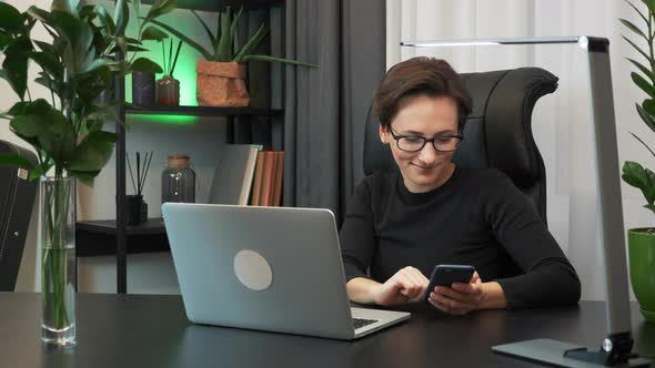 Woman is using smartphone sitting in modern office. Female works at workplace alt