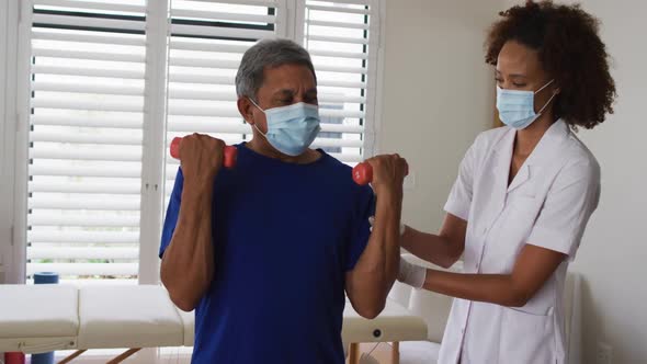 Mixed race female physiotherapist wearing mask helping senior exercise using dumbbells alt