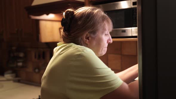 Eating disorder, hungry middle aged woman opens a refrigerator at night and eating unhealthy food. alt