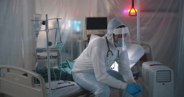 Sad Tired Doctor in Protective Suit and Medical Mask Sitting on Empty Bed in Hospital Ward alt