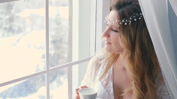 Young Slim Sexy Blond Girl in Lingerie and Boudoir Drinking a Tea and Looking in Window in Winter