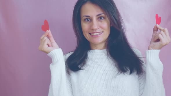 Smiling Funny Young Brunette in a White Sweater Smiles Blinks and Waving Red Hearts on a Pink
