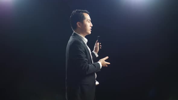 Side View Of Asian Speaker Man In Business Suit Holding And Pointing Smartphone While Speaking alt
