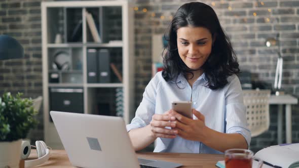 Pretty Girl Touching Smartphone Screen and Smiling Sitting at Desk in Office alt