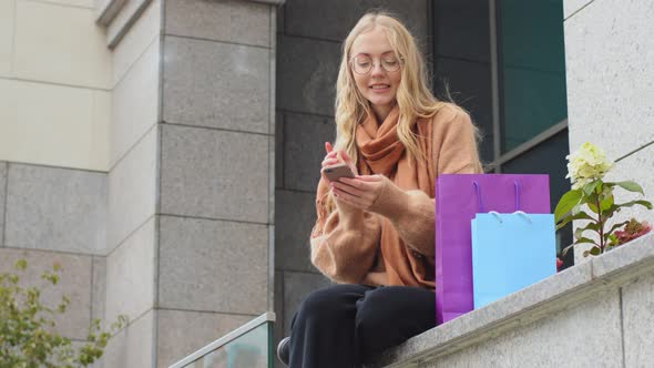 Female Shopper Sitting Outdoors Near Building with Colorful Bags Rejoices Discount Sale Black Friday alt