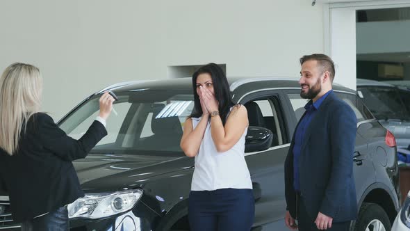 The Seller Hands Over the Keys To Your New Car Buyers. Girl Jumping of Joy and Hugs Her Husband