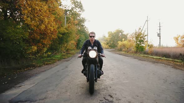 Front View of a Stylish Cool Young Man in Sunglasses and Leather Jacket Riding Motorcycle on a alt