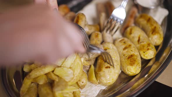 Closeup Top View Tray with Baked Grilled Fried Potato with Hands Pricking Sausages with Fork Leaving alt