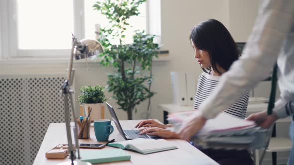 Young Woman Working with Laptop When Assistant Brigning Her Pile of Documents alt