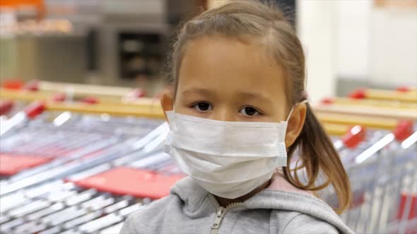 Portrait of Little Child Girl in Medical Mask Is Gazing at Camera in Supermarket