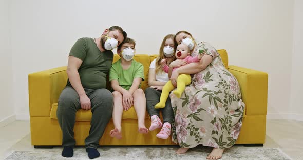 Family in Masks Sitting on Sofa. Overweight Mother and Father in Medical Masks Hugging Kids alt
