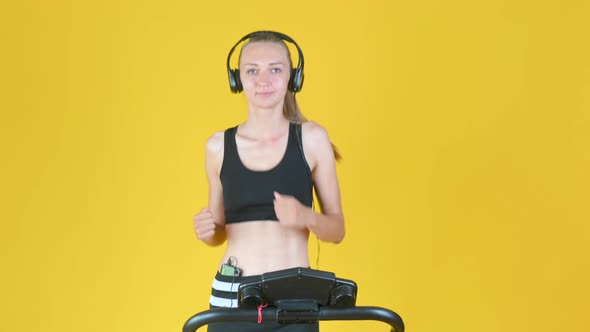 Fit blonde girl in sportswear with headphones trains on a treadmill. alt