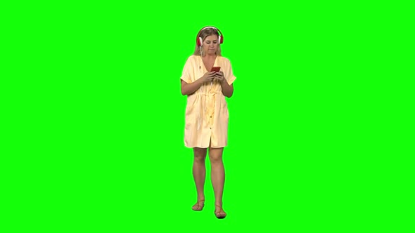 Tanned Blond Woman Walking in Big Red Headphones, Chooses Music on Mobile Phone on Greenscreen alt