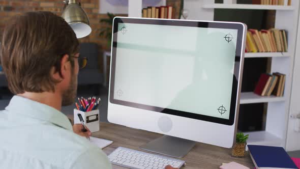 Caucasian male teacher taking notes while having a videocall on computer in the classroom at school alt