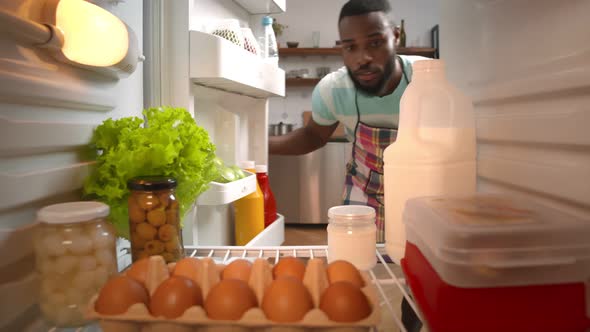 African Man in Apron Taking Fresh Ingredients From Fridge alt