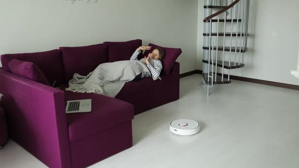 Free from household chores woman is relaxing on couch while robot vacuum cleaner cleanin.