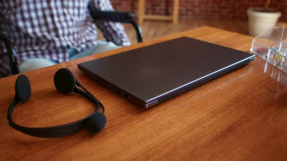 Disabled Man in Wheelchair Drives Up to Table Puts on Headphones and Opens Laptop Closeup of Table alt