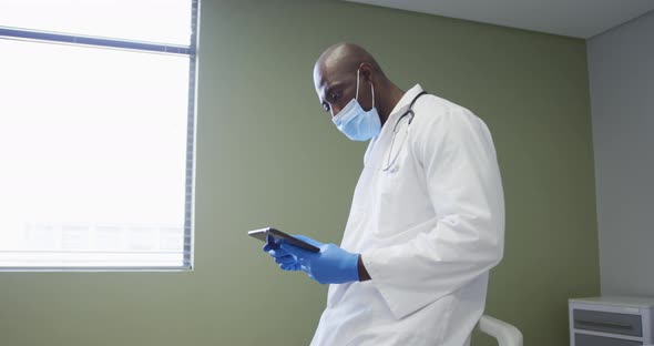 African american senior male doctor wearing face mask using digital tablet at hospital alt
