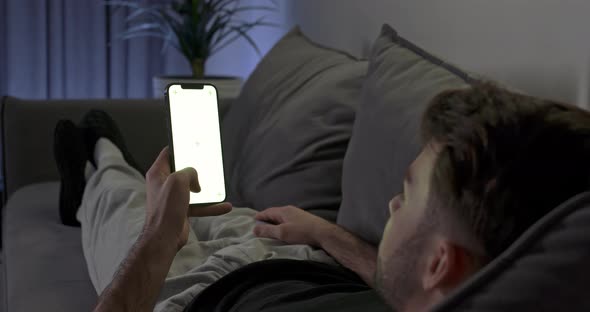 Guy Lying on the Sofa in Living Room Man Rests on the Sofa and Uses Smartphone Scrolls Through the alt