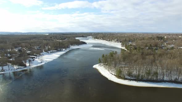 Aerial over icy river in winter snow on forested riverbanks alt