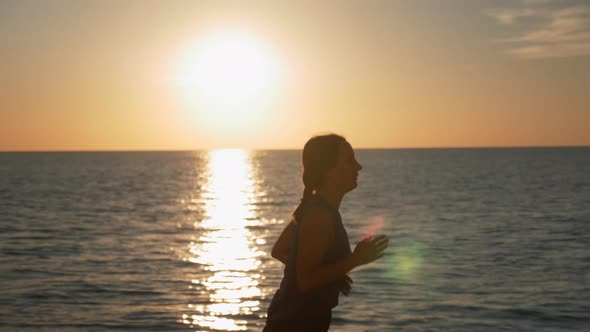 Concentrated brunette woman is running at sunrise and doing morning exercises on seashore. alt