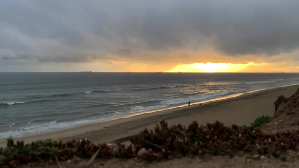 4k 60p, Golden sunset over ocean waves lapping onto a beach with a man walking his dog. Cargo ships alt