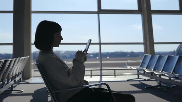 Young Woman Sitting at Airport and Using Modern Smartphone alt