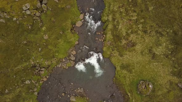 Aerial top down dolly out of river and Glymur waterfalls streaming down rocky cliff in verdant highl alt