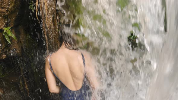 A Young Caucasian Girl in a Blue Swimsuit Walks Through a Large Wall of Falling Water on a Mountain