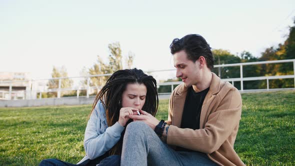 Happy Young Hipster Couple Sitting on Grass and Smoking Marijuana Joint in Park alt
