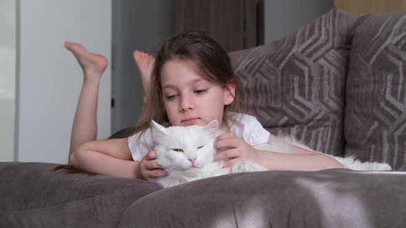 A Little Happy White Girl Lies on the Sofa in the Living Room Hugs a White Fluffy Cat and Smiles