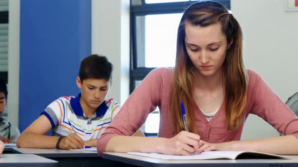 Schoolgirl passing chit to her classmate during exam in classroom alt