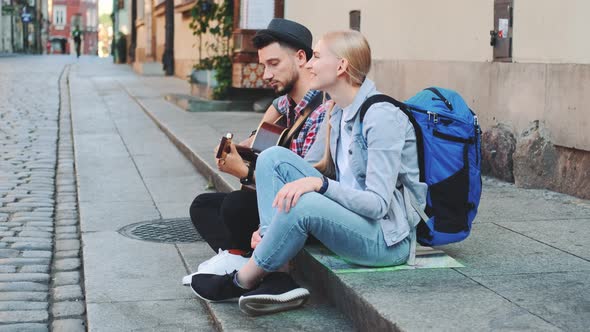 Couple of Tourists Sitting on Sidewalk, Playing Guitar and Having Rest alt