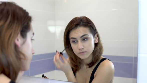 Young Woman Doing Eyelash Makeup. A Girl in a Black Bra in Front of a Bathroom Mirror. View Through alt