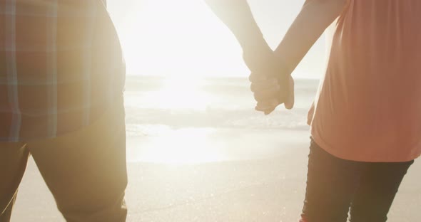 Senior african american couple holding hands and walking on sunny beach alt