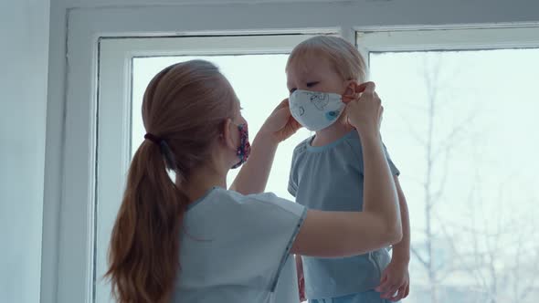 Mom with a Small Child in a Mask Are Sitting at the Window During Quarantine alt