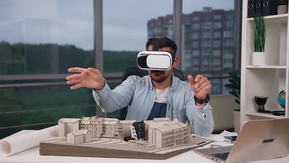 Designer Sitting at His Workplace with Constructed Model of New Buildings and Working on VR alt