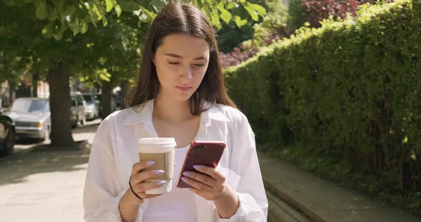 Young Woman with Take Away Cup and Drink Hot Coffee, Looking at Smartphone, Dressed in Business Suit alt