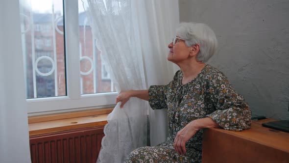 Upset Caucasian Pensioner in Her 70s Sitting Alone Next to the Window and Looking Out of the Window alt
