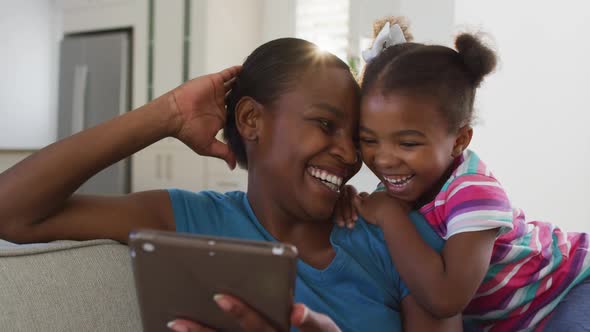 Happy african american mother and daughter sitting on sofa using digital tablet and laughing alt