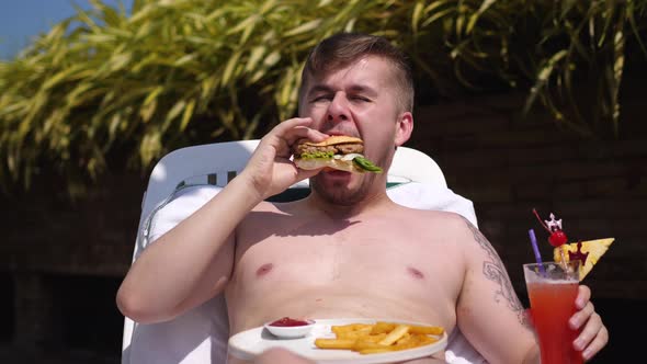 Bearded Man Is Eating a Hamburger Lying on a Lounger with a Cocktail
