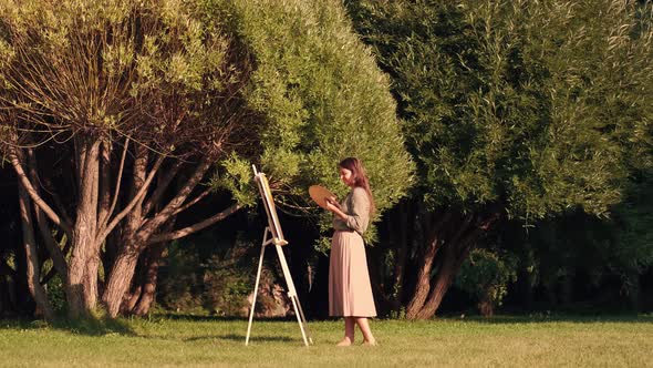 Cheerful Woman Artist Paints a Picture on Canvas While Standing in a Park in Sunny Weather alt