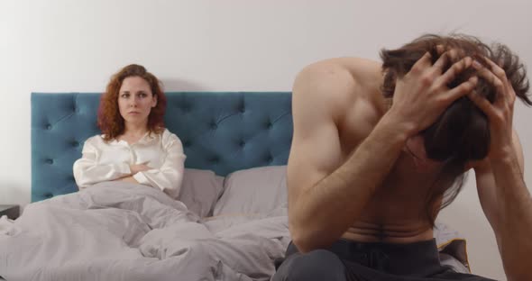 Worried Man in Tension Sitting at Bed with Upset Woman Lying on Background alt