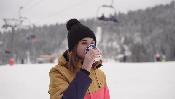 Girl with Takeaway Coffee Cup Over Ski Resort Background is Drinking and Waving Someone alt