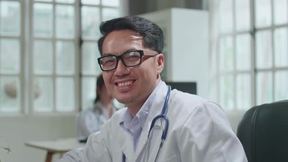 Asian Man Doctor Is Using Desktop Computer, He Turns And Smiles To Camera In Workplace alt