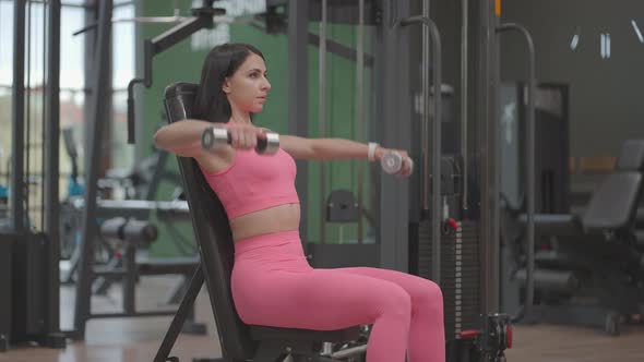A Brunette Woman in a Pink Suit Raises Dumbbells to the Sides While Training Her Shoulders in the alt