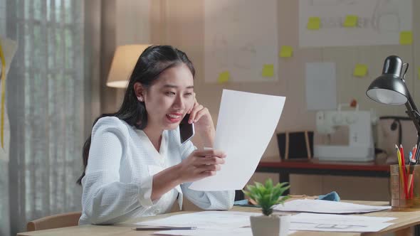 Asian Woman Designer Talking On Smartphone While Looking At The Layout Bond In Hand At The Office alt