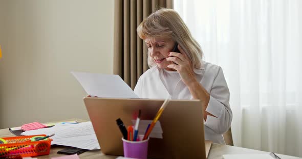Senior Woman Grandmother Works Online at Home Sits at Desk and Laptop Talks on the Phone Solves alt